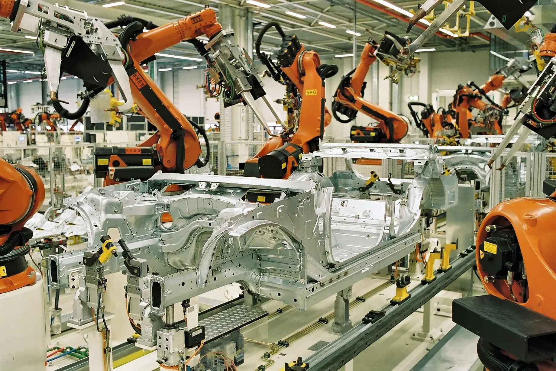 Orange industrial robots spot-welding bare car bodies on a production line.