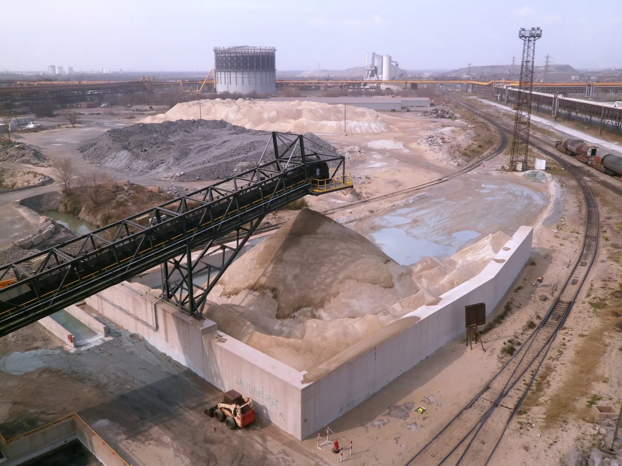 Heaps of pale granulated and grey crystallised blast-furnace slag at a steelworks, with a conveyor, rail tracks and a gas holder.