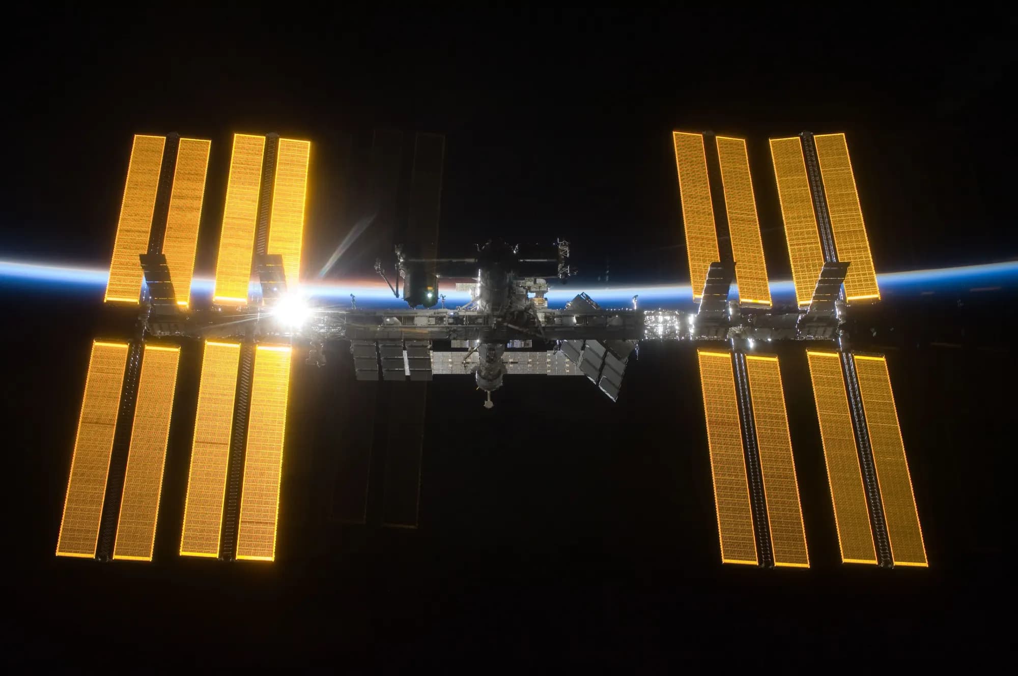 The International Space Station with its golden solar arrays, seen against black space and the thin blue line of Earth's atmosphere, with the Sun behind it.