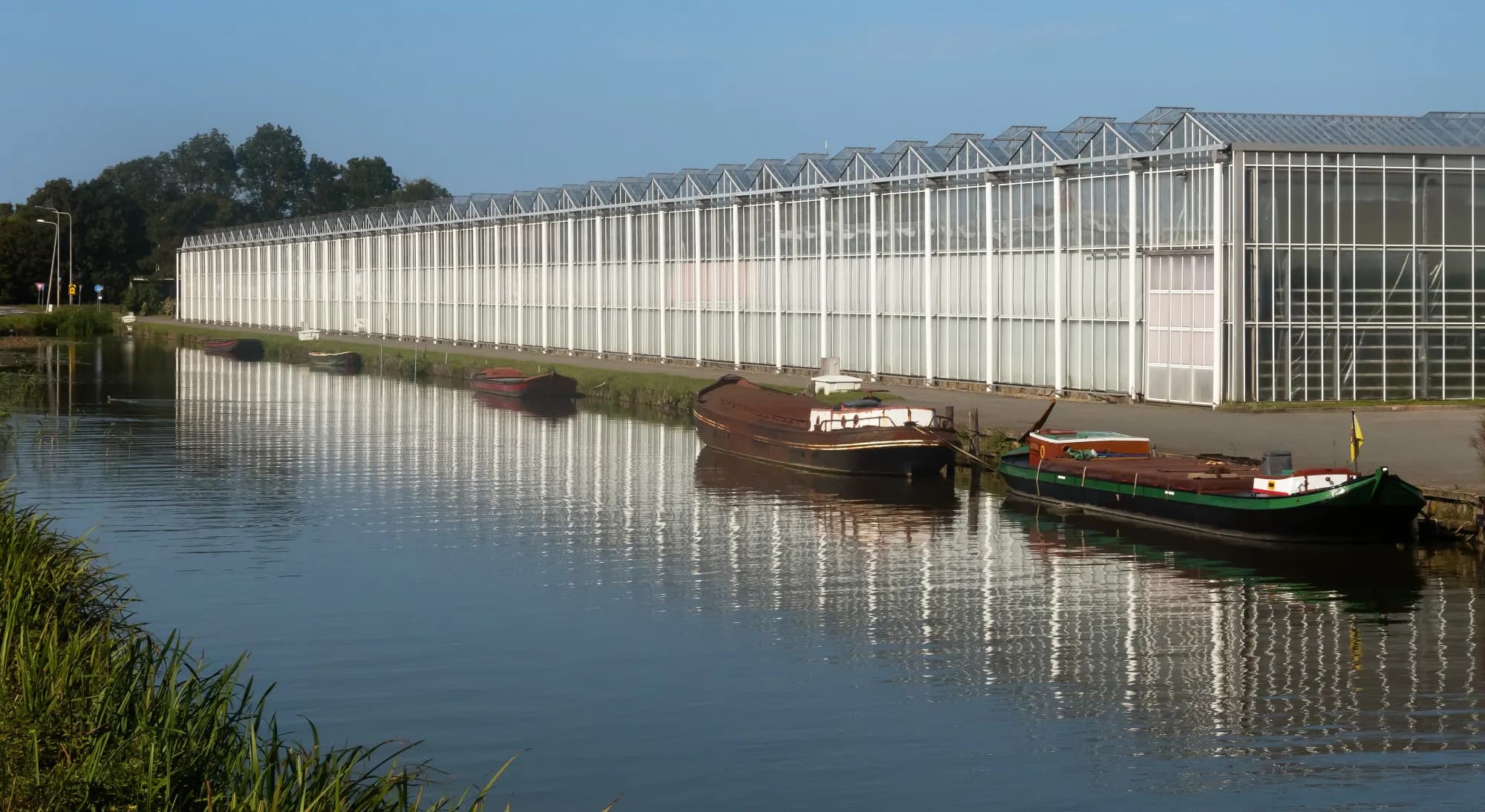 A long glass greenhouse along a canal at Poeldijk in the Westland, reflected in the still water with moored boats.