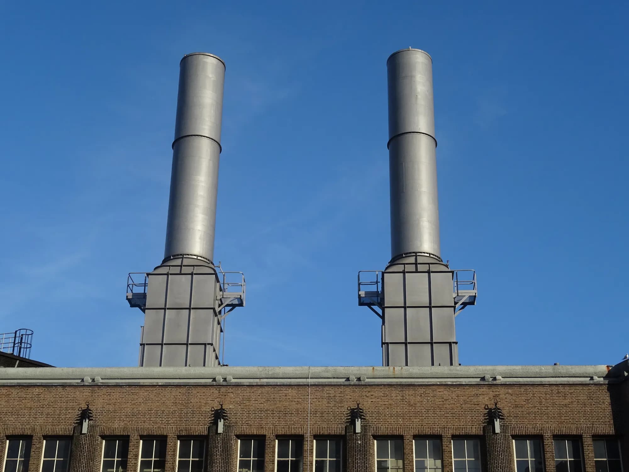 Two steel chimneys rising above the brick facade of a district heating station against a deep blue sky.